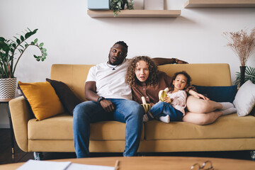 Diverse family resting on sofa and looking at camera in shocking surprise