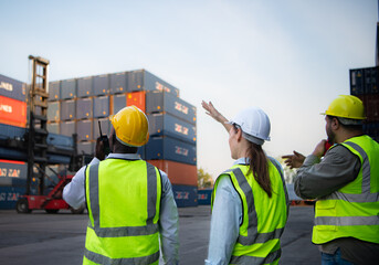 Group of workers in the import and export industry use walkie talkies to communicate with drivers of reach stacker containers in an empty container warehouse.