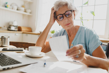 Indoor image of thoughtful puzzled senior woman with gray hair in glasses looking at paycheck holding head in hand, upset with high rent charge, sitting at kitchen table in front of laptop - obrazy, fototapety, plakaty