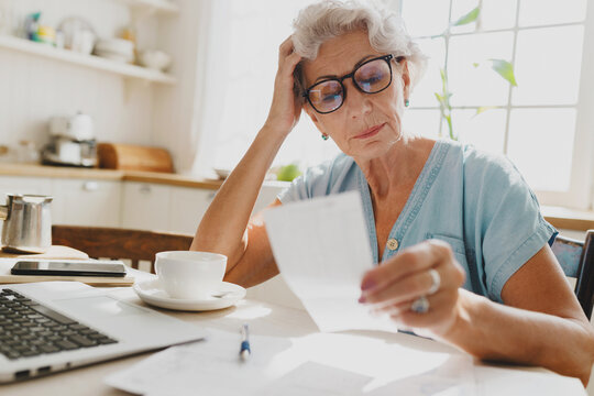 Indoor image of thoughtful puzzled senior woman with gray hair in glasses looking at paycheck holding head in hand, upset with high rent charge, sitting at kitchen table in front of laptop