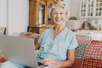 Joyful elderly lady in blue overalls using laptop on sofa in lovely living room, shopping online,...