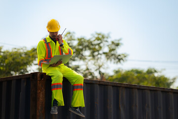 Workers in the import and export industry holding a laptop and sitting on a shipping container to inspect the container's cargo.