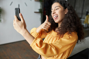 Side view of funny black girl of 20s recording short video or taking selfie on smartphone grimacing, making faces, sticking tongue and showing thumb up, having fun being in good mood - obrazy, fototapety, plakaty
