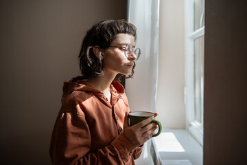 Teen girl nerd in glasses looking at window drinking cup of tea at home. Serious young woman...