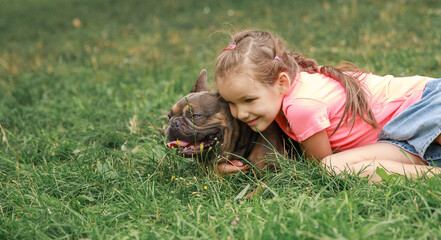 Bulldog lies in the grass. A girl plays with a dog. A child strokes an animal. The dog is resting in the cool greenery. Pet on a walk.
