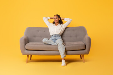 Relaxed black teenager girl resting sitting on couch, yellow backdrop