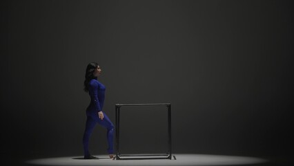 Fototapeta premium Portrait of female acrobat isolated on black background. Woman gymnastic dancer in blue sparkling bodysuit showing element on a cube.