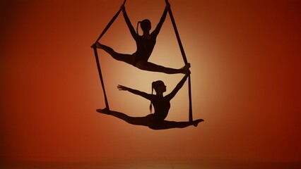 Silhouette of two female acrobats isolated on orange neon background. Girls aerial dancers performing acrobatic splits on ropes. © kinomaster
