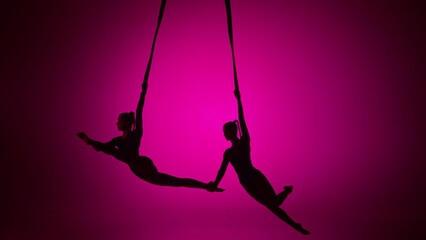 Silhouette of two female acrobats isolated on pink neon background. Girls aerial dancers performing flying element on ropes.