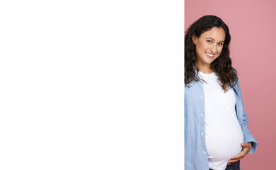 Beautiful young pregnant woman standing by white blank board