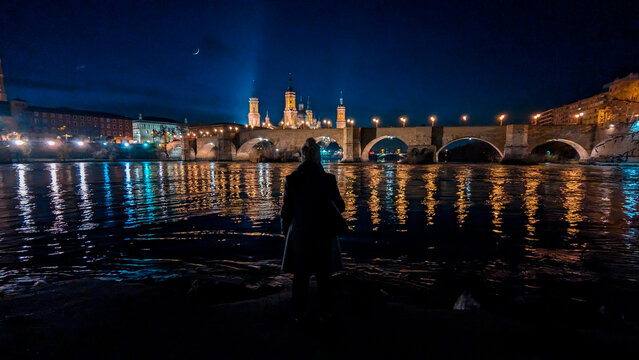 River of Reflections: A woman's backlit beauty harmonizes with Zaragoza's night, a vision by the Ebro.
