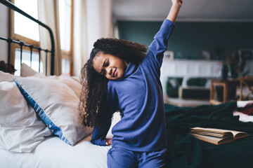 Positive black girl raising hand while waking up from bed in bedroom