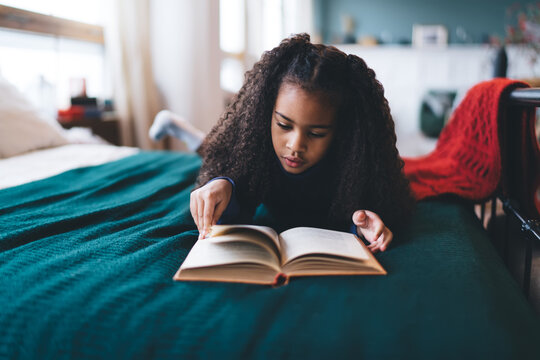 Focused black girl reading book while lying on bed at home