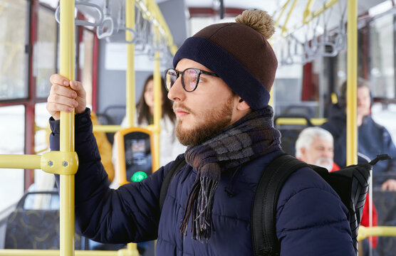 Close up of pensive adult bearded male in glasses with backpack standing looking at window in transport. Crop of young man and blur passengers sitting behind in moving bus. Concept of city life.