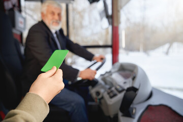 Close up view of hand holding green payment card showing to driver while boarding in public transport. Crop of hand with paying ticket in front of blurred driver. Concept of transportation.