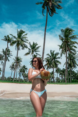Young beautiful fit female model pose at exotic beach in a white bikini with coconut in the hands