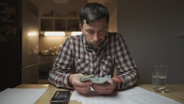Frustrated man looking at bills and calculating money on calculator in kitchen apartment. Depressed male checking finance. Cash issues. Counting invoices and tax expenditures. Public service billing. 