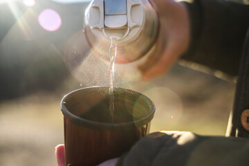 Woman is pouring hot drink from thermos into travel mug, close-up, warming drink in cold weather, active leisure, A hiker pours hot tea into a cup on a cold autumn or winter day