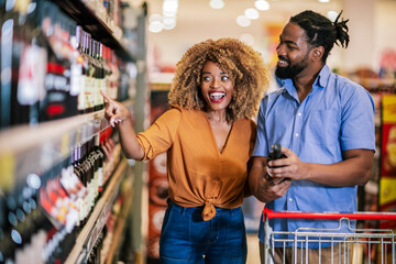 African American couple with trolley purchasing groceries at mall. Buying wine. - obrazy, fototapety, plakaty