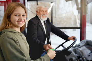 Close up of young female in olive hoodie and elderly driver together holding ticket near driver place in public transport. Crop of cute girl paying for trip in bus. Concept of city transportation.