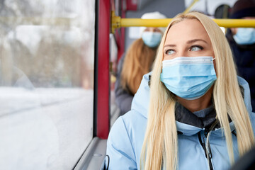 Fototapeta na wymiar Front view of pretty female wearing protective mask and blue jacket sitting with passengers behind in bus. Close up of adult woman looking pensive at window in moving transport. Concept of city life.