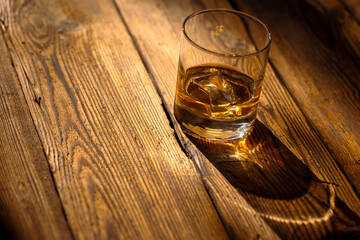 Glass with whisky with ice on a textured wooden table in harsh sunlight.