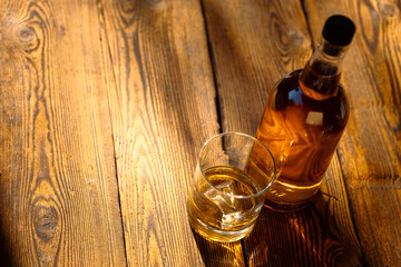 Bottle of whisky with glass and ice on a textured aged wooden table in harsh sunlight.