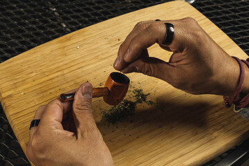 Man preparing his pipe with marijuana on a wooden board, with remains of ground marijuana around.
