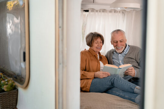 Happy senior elderly old couple reading book, watching album with photos for memories while traveling together in camper van trailer motor home. Eco-tourism concept