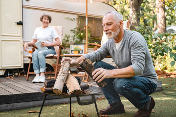 Careful senior old elderly husband laying a fire with firewood while wife relaxing on the porch of...