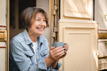 Smiling relaxed old senior elderly traveler woman grandmother drinking coffee resting on the porch of trailer camper van motor wheel home - obrazy, fototapety, plakaty