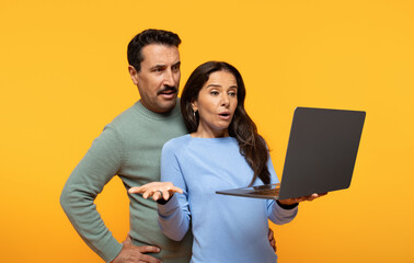 Surprised man and woman in casual attire react to shocking news on a laptop screen with incredulous...