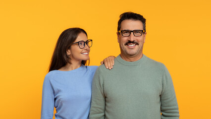 A happy couple stands shoulder to shoulder, the woman wearing glasses