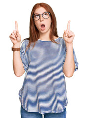 Young read head woman wearing casual clothes and glasses amazed and surprised looking up and...