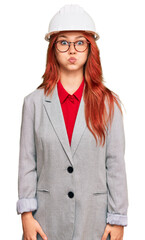 Young redhead woman wearing architect hardhat puffing cheeks with funny face. mouth inflated with air, crazy expression.