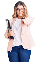 Young caucasian woman wearing business clothes and glasses holding binder pointing with finger to the camera and to you, confident gesture looking serious