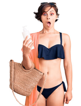 Beautiful young woman with short hair wearing bikini holding sunscreen lotion scared and amazed with open mouth for surprise, disbelief face