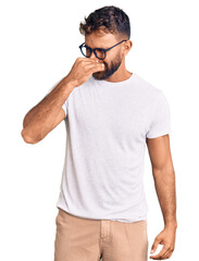 Young hispanic man wearing casual clothes and glasses smelling something stinky and disgusting,...