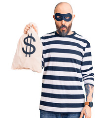 Young handsome man wearing burglar mask holding money bag with dollar symbol scared and amazed with...