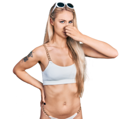 Beautiful young blonde woman wearing bikini smelling something stinky and disgusting, intolerable smell, holding breath with fingers on nose. bad smell