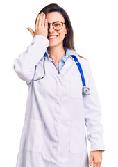 Young beautiful woman wearing doctor stethoscope and glasses covering one eye with hand, confident smile on face and surprise emotion.