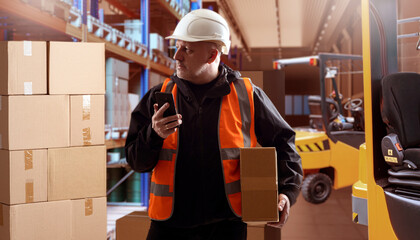 Man warehouse contractor. Factory storage manager. Product storage area. Clipboard man thought. Warehouse contractor inspects goods. Tiered racks with boxes behind guy. Logistics, fulfillment - obrazy, fototapety, plakaty
