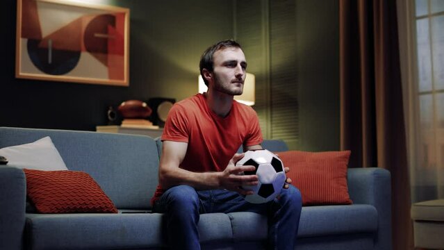 Male observing football game. Concentrated man holding pillow in shape of soccer ball. Male watching football match. Man sad for fail of favorite team. Male worrying for final match.