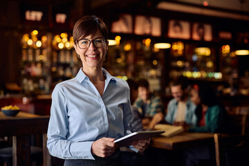 Happy waitress using touchpad while working in bar and looking at camera.