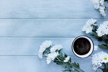 White enamel cup of hot steaming coffee surrounded by white Chrysanthemum flowers over a blue rustic table. Table top view.
