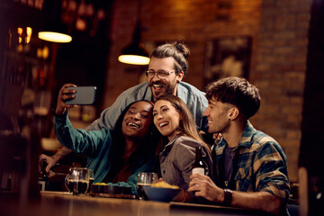 Multiracial group of friends taking selfie with cell phone in pub.