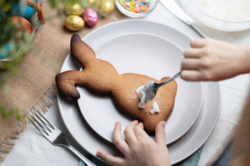 Close-up of bunny shaped gingerbread cookie. Hands of child decorating biscuit with icing on plate. Easter holiday traditions, family time