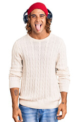 Young hispanic man listening to music using headphones sticking tongue out happy with funny expression. emotion concept.