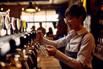 Happy female barista pouring lager beer from beer tap at bar counter.