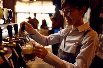 Fototapeta na wymiar Happy female barista pouring beer from beer tap while working in pub.